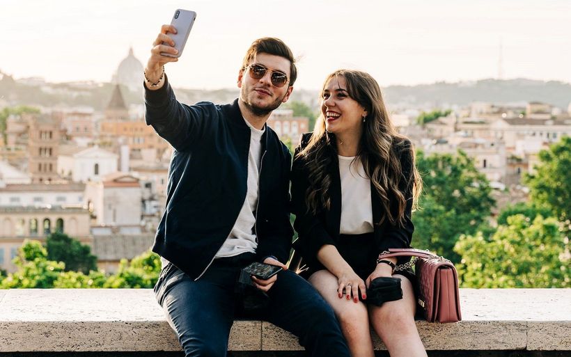 Ein junger Mann und eine junge Frau hocken auf einer Mauer und machen ein Selfie.